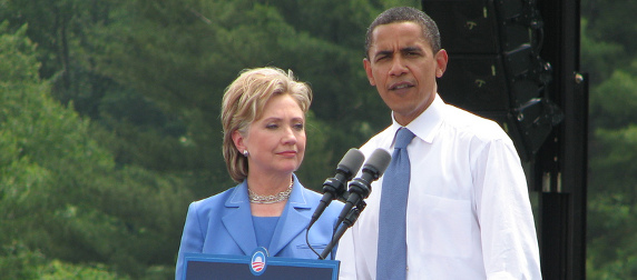 Barack Obama speaks at a podium while Hillary Clinton stands at his side