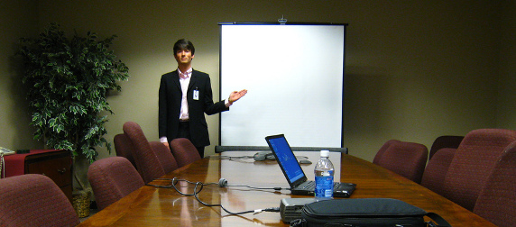 man in a suit standing at the end of a conference table, indicating a black projector screen