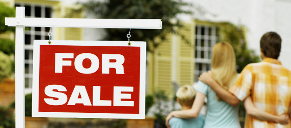 Red 'For Sale' sign, with man, woman and child looking at a house in a blurred background