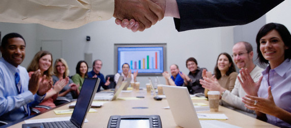 men and women in business attire seated at a conference table applaud a handshake in the foreground