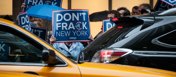 protestor holding a sign reading Don't Frack New York in front of face, viewed across traffic