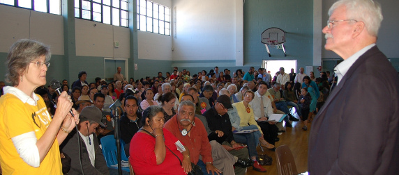 George Miller addressed by woman in yellow shirt with microphone at a town hall meeting