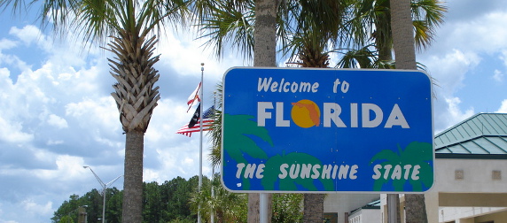 Welcome to Florida sign in front of palm trees, flags and a blue sky