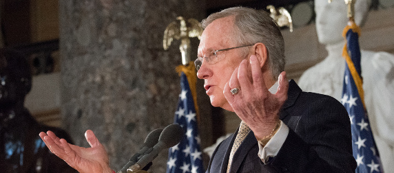 Harry Reid speaking at a podium, two American flags in the background