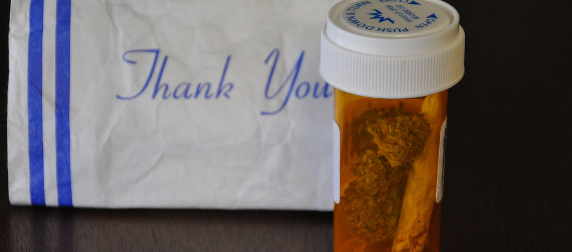 medical cannibis in an orange prescription bottle, in front of a white paper bag that says Thank You