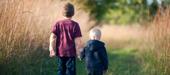 two children holding hands, walking away from the camera on a path through a wheat field