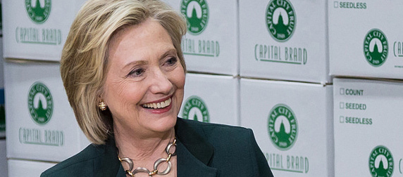 Hillary Clinton, smiling, in front of white boxes labeled 'Capital Brand' with a green logo