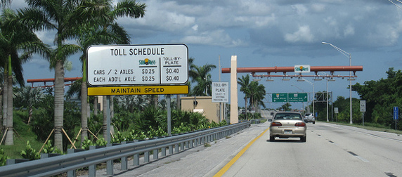 toll schedule by the side of a two-lane road, listing rates for SunPass holders and toll-by-plate drivers