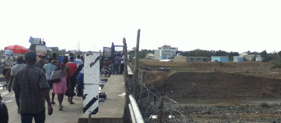 people walking over a bridge lined with curled barbed wire, dirt hills on the other side