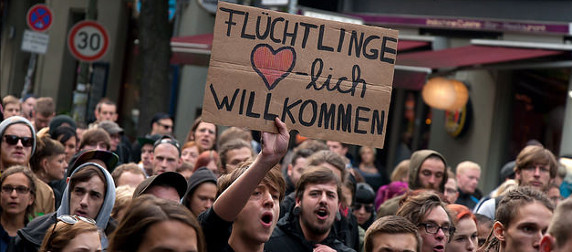 protestors, one holding a cardboard sign that reads 'fluchtlinge herzlich willkommen'