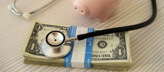 stethoscope resting on a stack of $1 bills, next to a piggy bank
