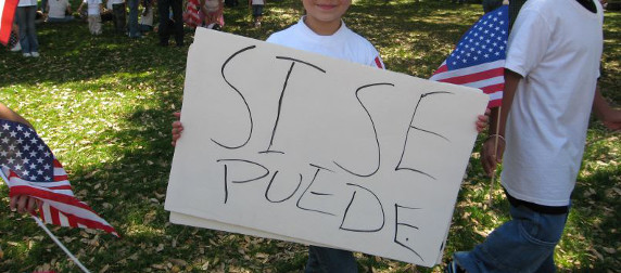 hand-drawn poster reading SI SE PUEDE, held by a child, with American flags in the background