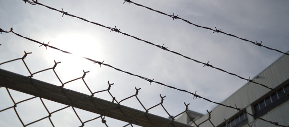 chain-link fence, topped with barbed wire, against an industrial style building and sunny sky