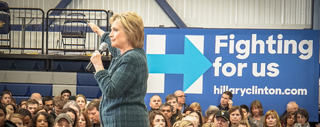 Hillary Clinton speaking to a crowd in front of a banner that reads Fighting For Us hillaryclinton.com