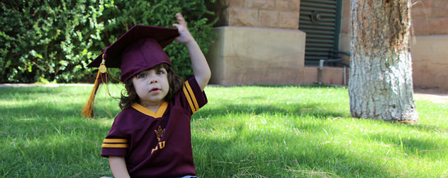 toddler wearing a mortarboard and ASU jersey, seated on grass