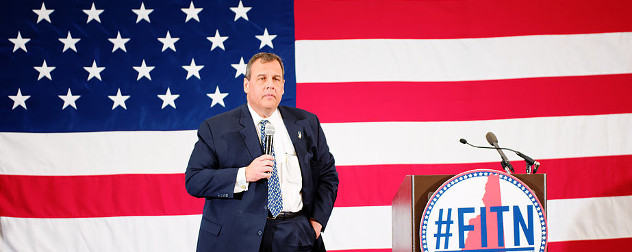 Chris Christie with a microphone in front of an American flag, a podium with the banner #FITN on his left