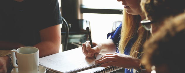 details of person with long hair, person with glasses and person wearing black t-shirt working at a table with a notebook and coffee mug