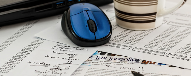 pile of tax documents with a computer mouse, coffee mug and pen on top