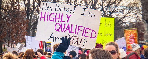 protester holding a sign that reads 'Hey Betsy! I'm highly qualified - are you?'