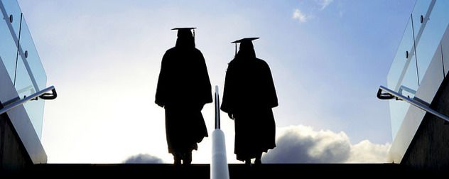 two people in graduation cap and gown, seen in silhouette climbing a staircase toward a blue sky
