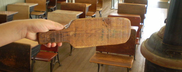 wooden paddle held out in front of an empty, old-fashioned school classroom