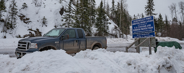U.S.-Canada border crossing warning sign in a snowy scene with a pickup truck