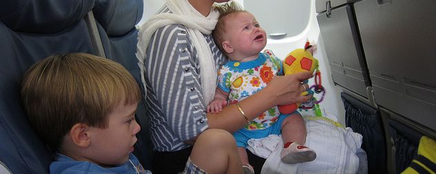 upset toddler in an adult's lap on an airline seat, with a slouching child in the adjacent seat