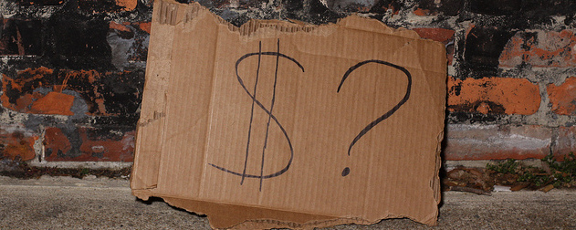 cardboard sign, possibly from homeless person, with the simple message '$?' propped against a brick wall.