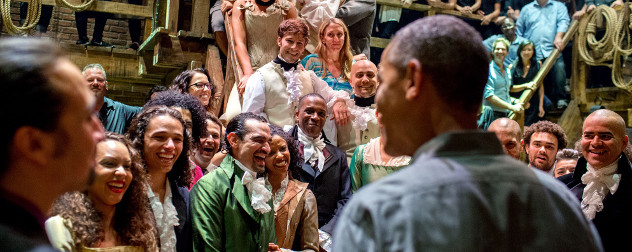 Barack Obama, shot from behind, interacting with the original cast and crew of the Broadway musical 'Hamilton' on the show's set.