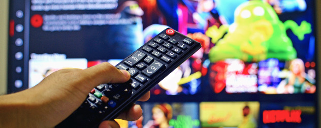 hand pointing a remote control at an out-of-focus TV screen with a streaming video service player menu displayed.