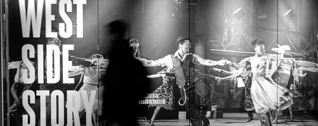 black and white image of a person walking past an outdoor ad for 'West Side Story.'