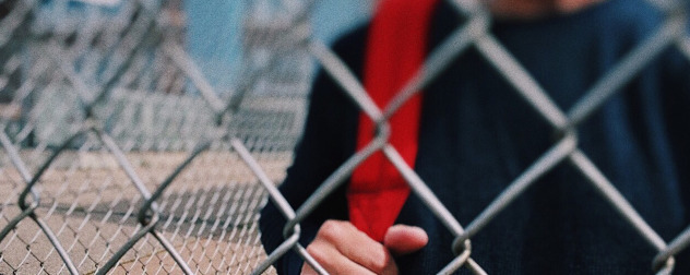 detail of student with red book bag strap behind chain-link fence, presumably at school.