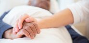 young person's hand on an old person's hand resting on a bed.