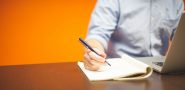 person holding a pen over an open notebook while working on a laptop (detail).