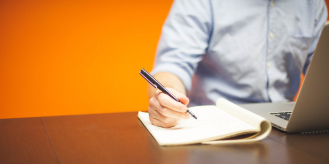 person holding a pen over an open notebook while working on a laptop (detail).